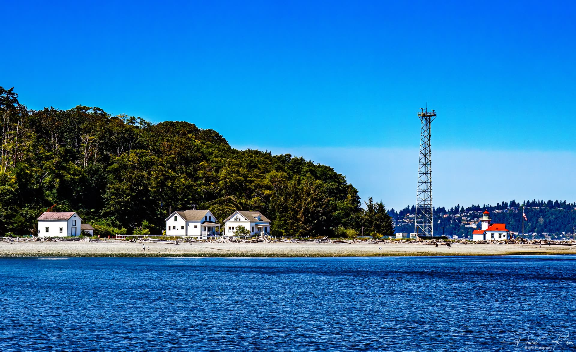 Shoreline scene with trees, waterfront buildings, and a tall communications structure.